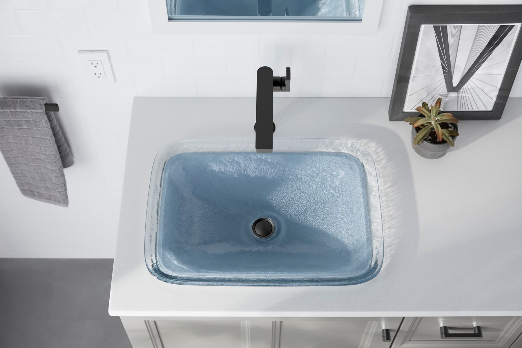 An overhead view of a black bathroom sink faucet and a glass bathroom sink with blue tint, backed by a white wall