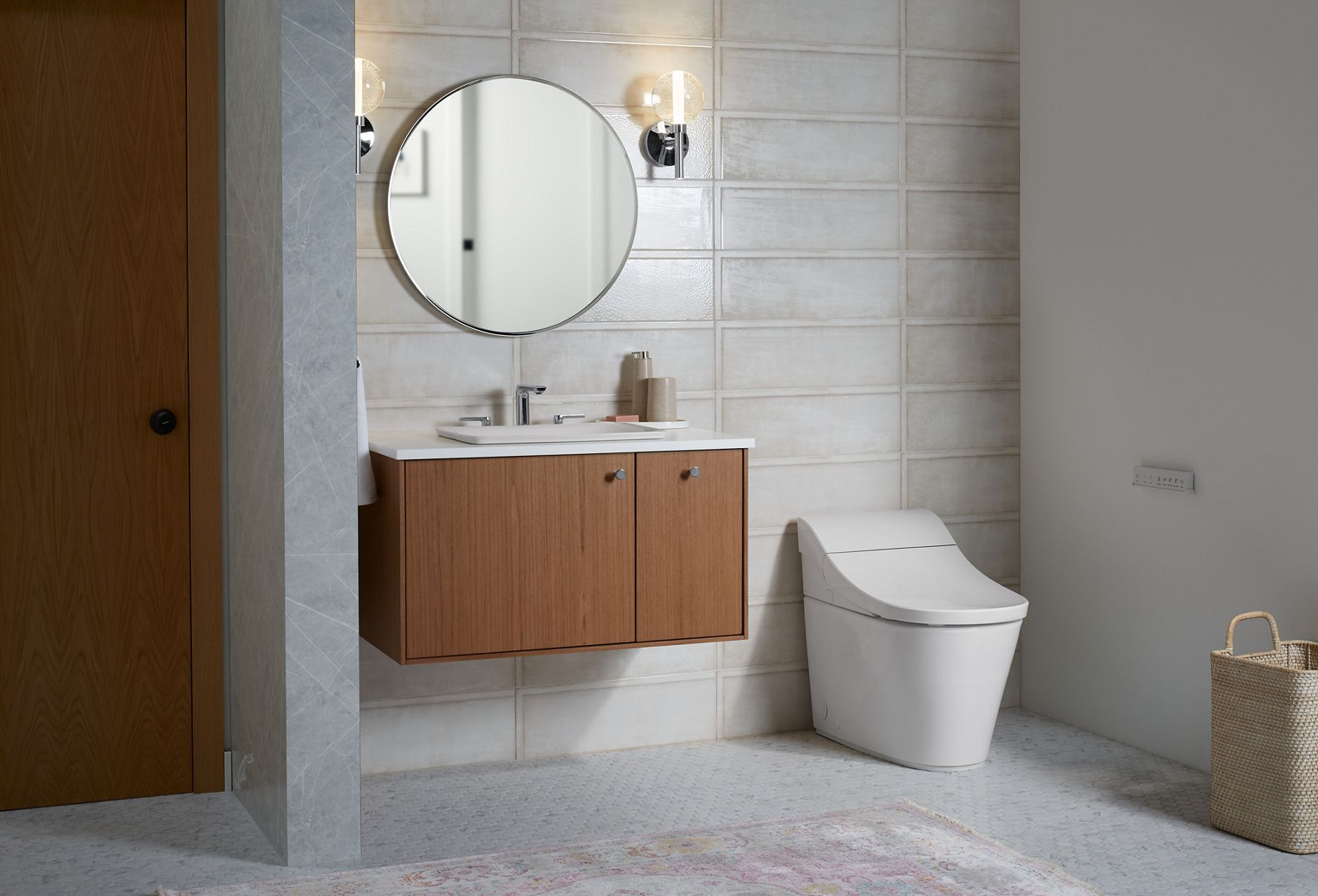 A white Innate toilet on a mottled white tile floor with a wall behind it consisting of large scale rectangular white tiles with weathered gray edges.