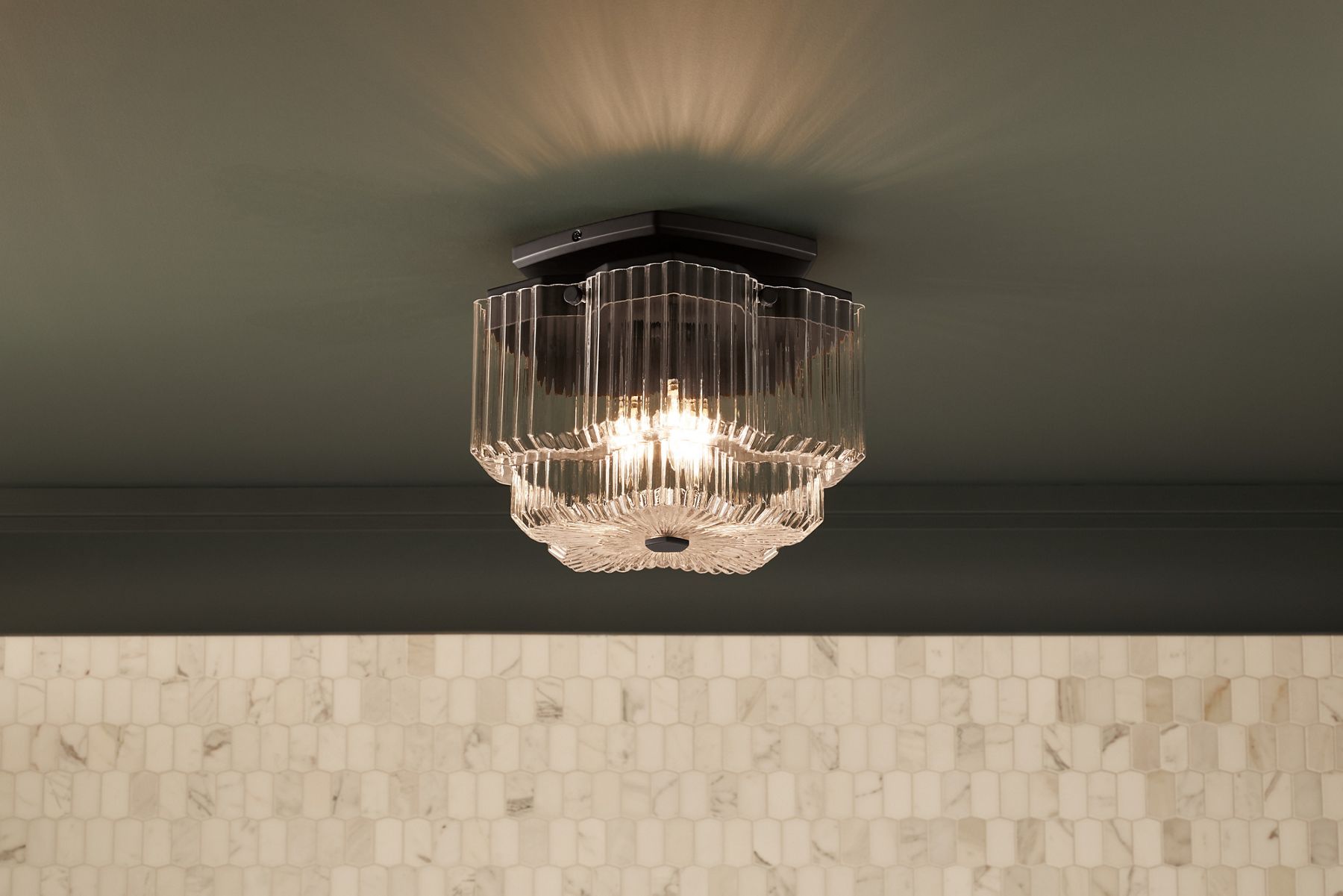 An Occasion three-light flushmount light on a dark green ceiling with a wall in cream-colored marble tile.