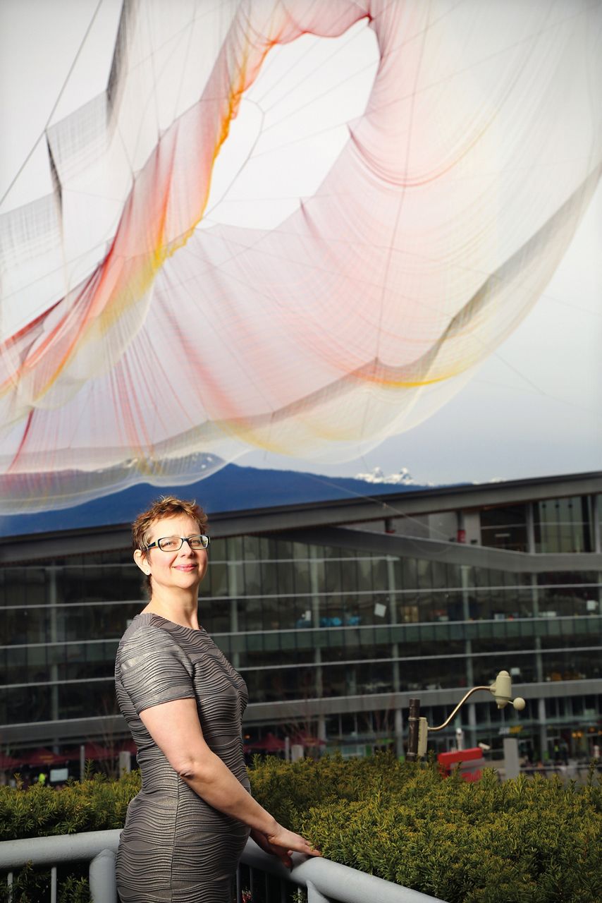 Janet Echelman posant sous une partie de sa grande installation artistique de filets roses et orange. Son corps est tourné vers la droite de l’image et sa tête est tournée vers l’appareil photo. Elle et son installation artistique sont éclairées par la gauche.