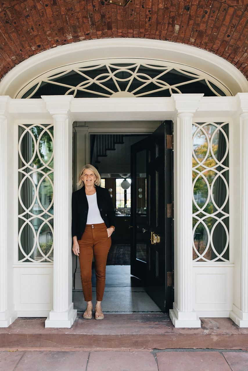 Designer Jean Stoffer wearing brown pants, a white shirt and a dark blue blazer leaning against the open door way to the home she designed.