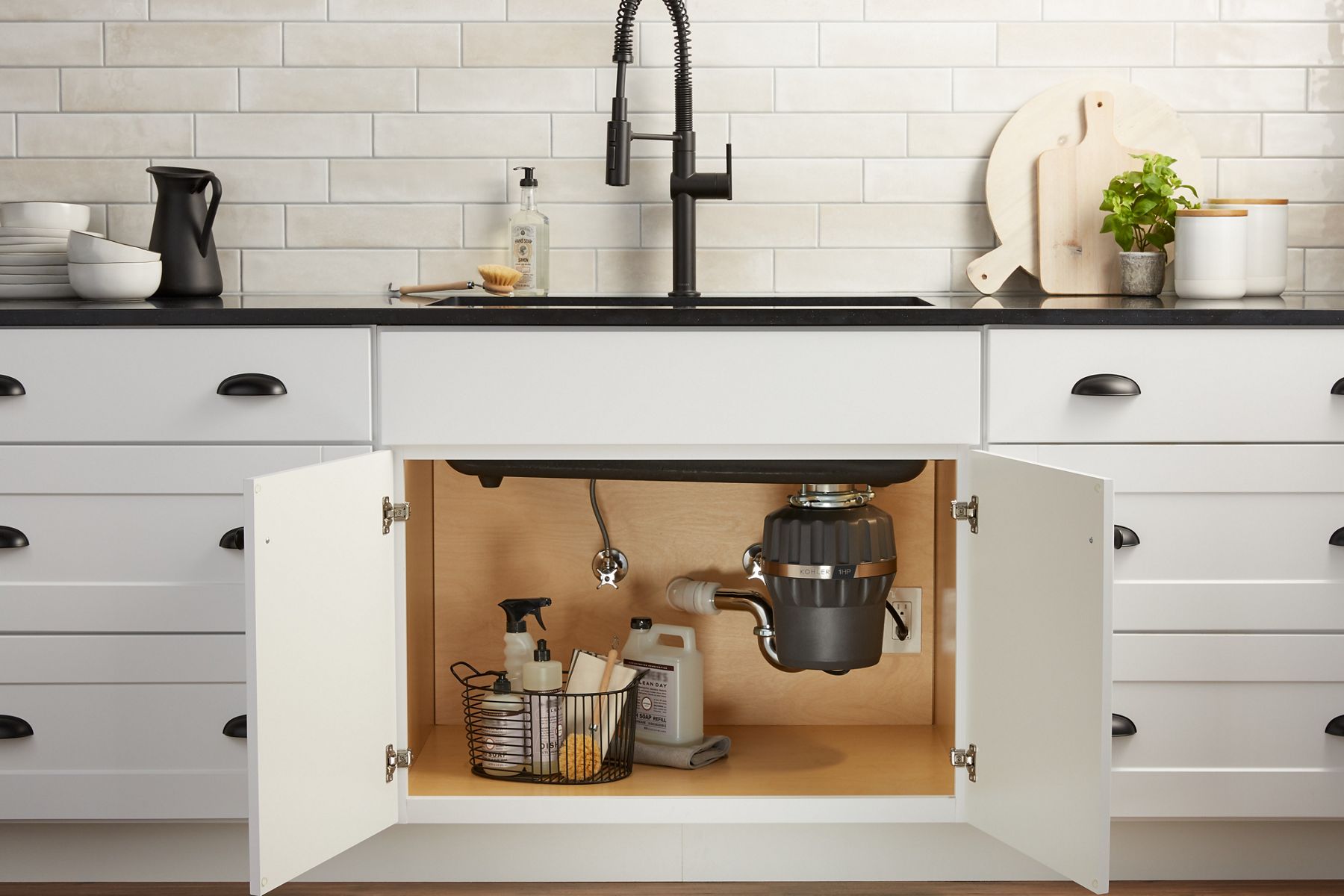 A white kitchen cabinet with the door under the sink opened up to show a garbage disposal.