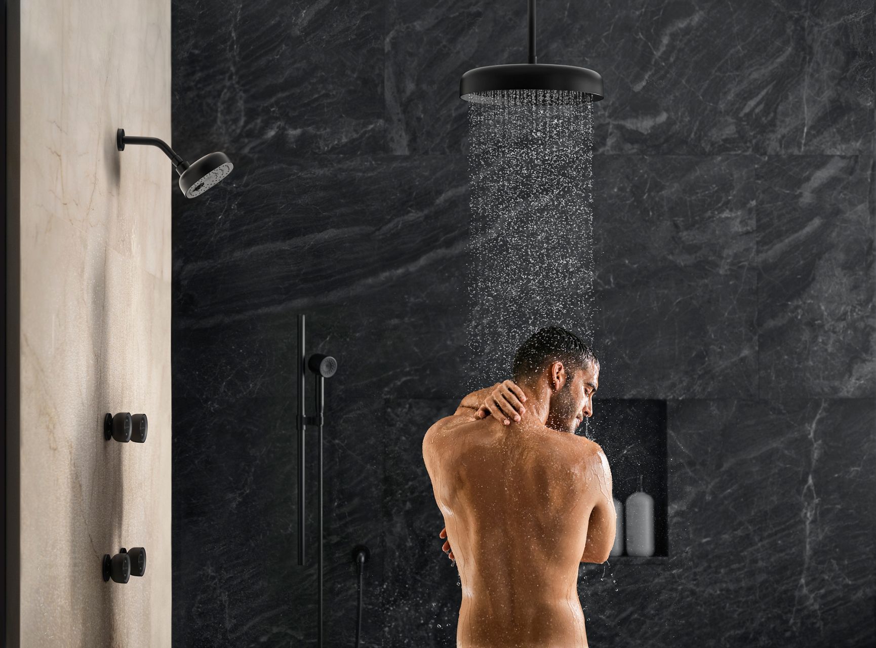 A man standing under a rainhead in a shower with gray stone walls.