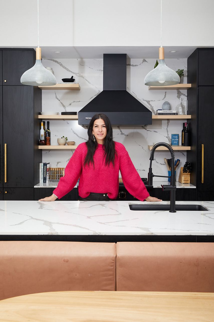 La créatrice de mode Rebecca Minkoff, vêtue d’un chandail à manches longues rose vif et d’un pantalon noir, debout derrière l’îlot de sa cuisine, qui présente des armoires gris foncé avec des ferrures en laiton brossé, un comptoir et un dosseret en marbre, et un robinet KOHLER Crue noir mat au premier plan.
