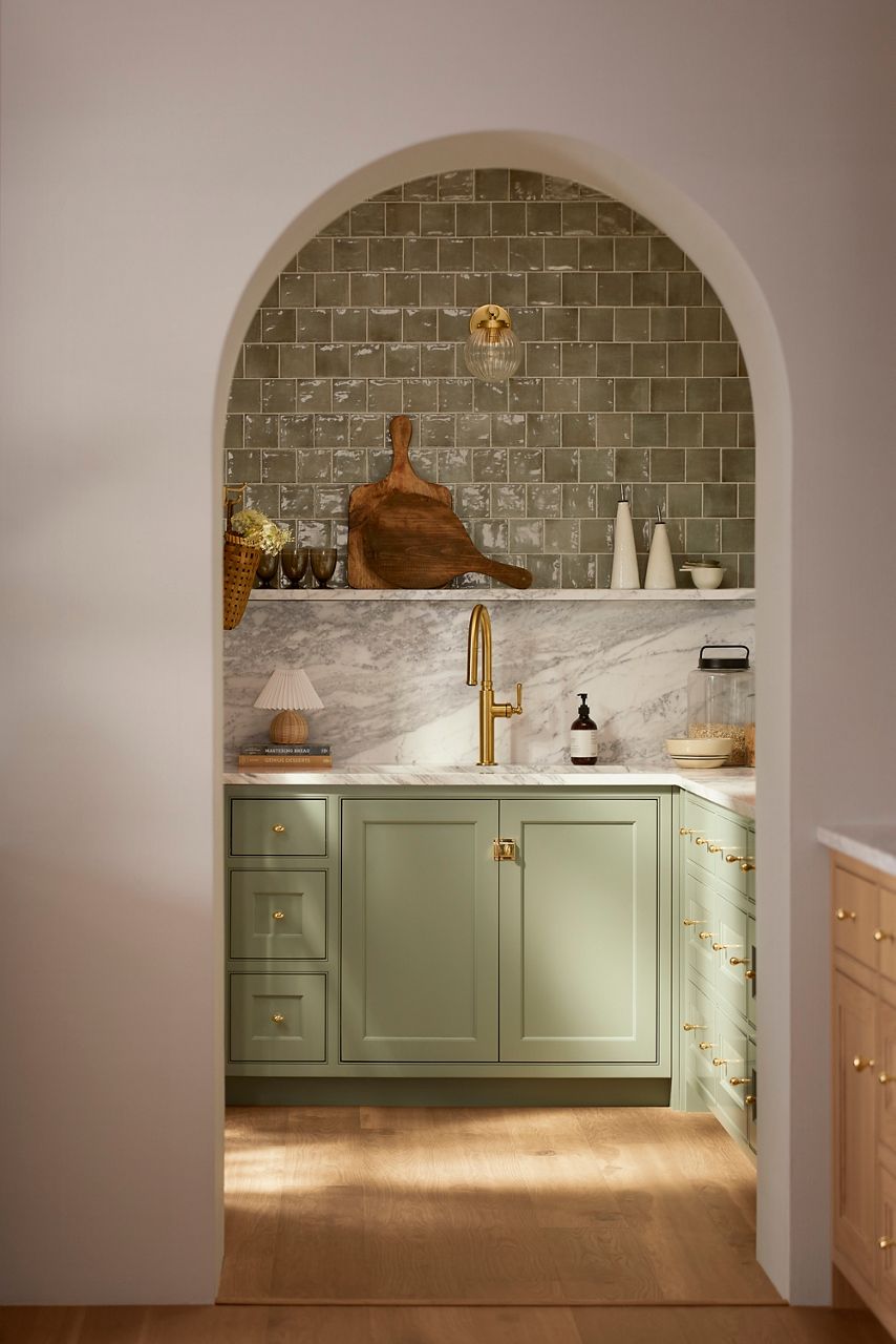A modern kitchen with rustic elements featuring a gold faucet, sage green cabinets, and wood cutting boards.