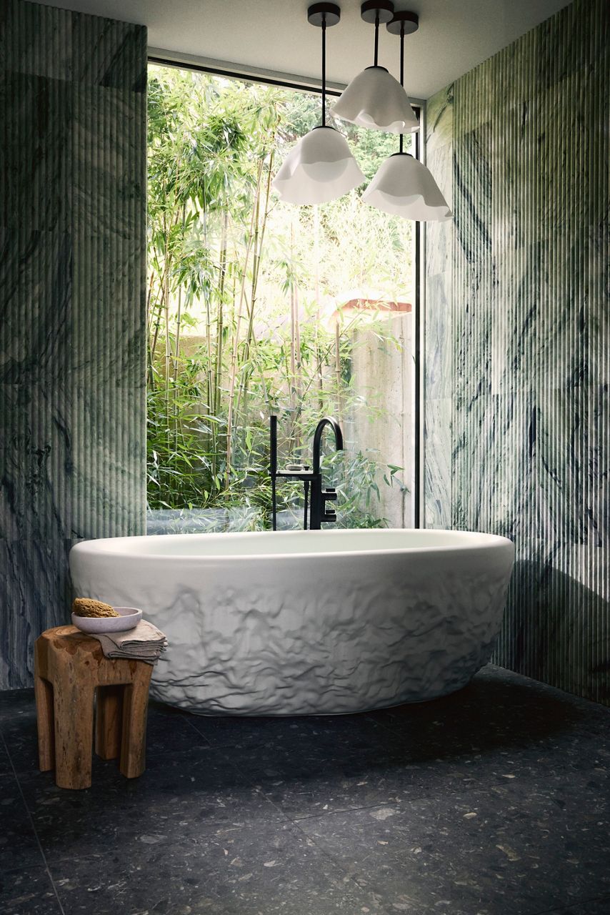 A white freestanding bathtub with textural base and black bathtub filler, backed by a window filled with greenery and a wooden stool and accessories in the foreground.