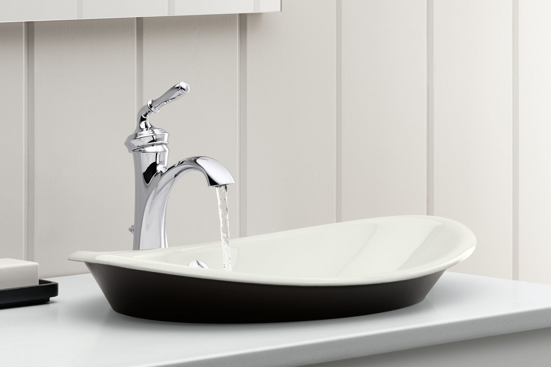 A close-up of a chrome bathroom sink faucet with running water over an enameled cast iron bathoom sink with white bowl and black base