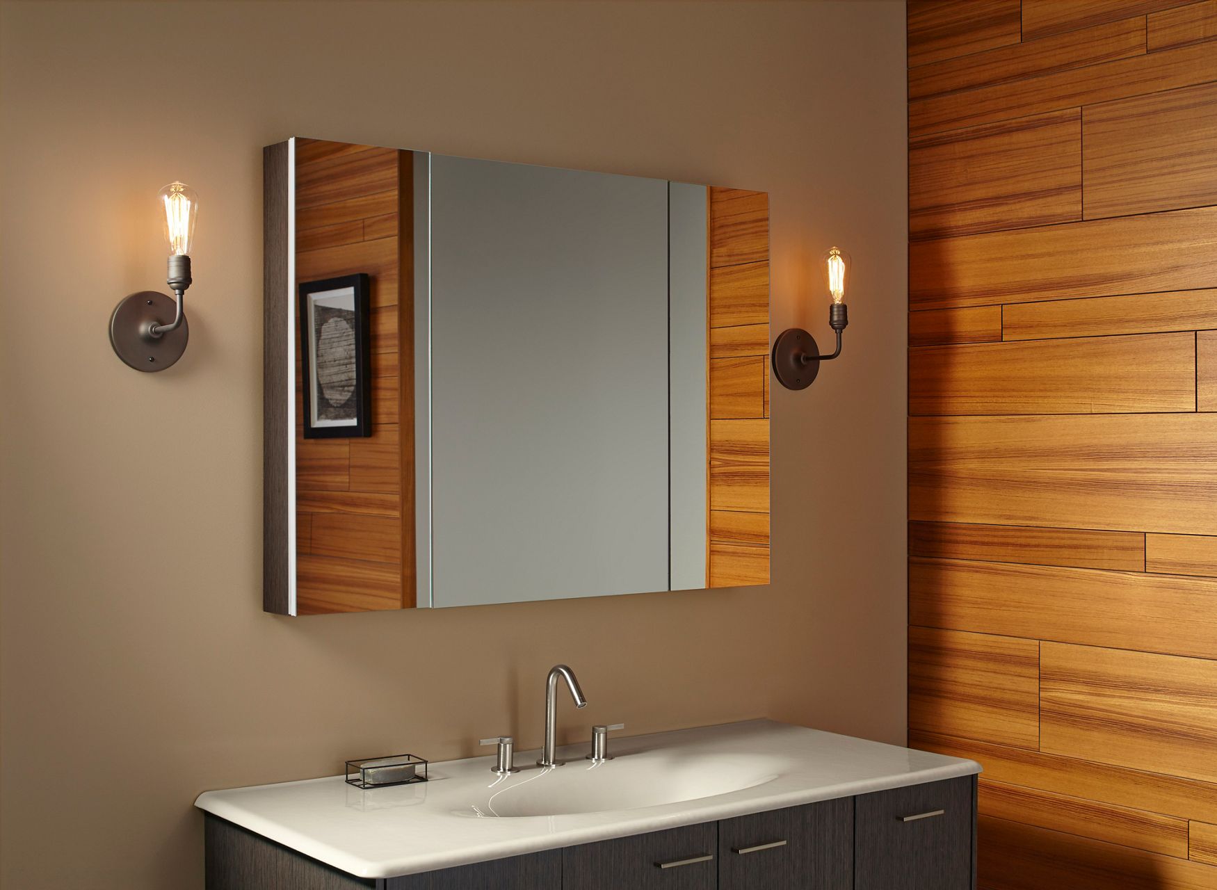 A large three-door medicine cabinet over a bathroom vanity sink and faucet with a illuminated wall sconce on one side