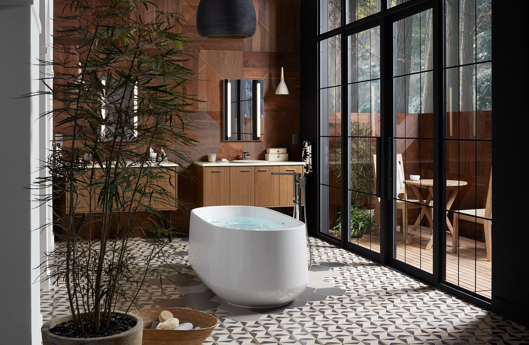 A white freestanding bath on a geometric tile floor in a light-filled bathroom with large glass doors on the right and two vanity spaces in the background.