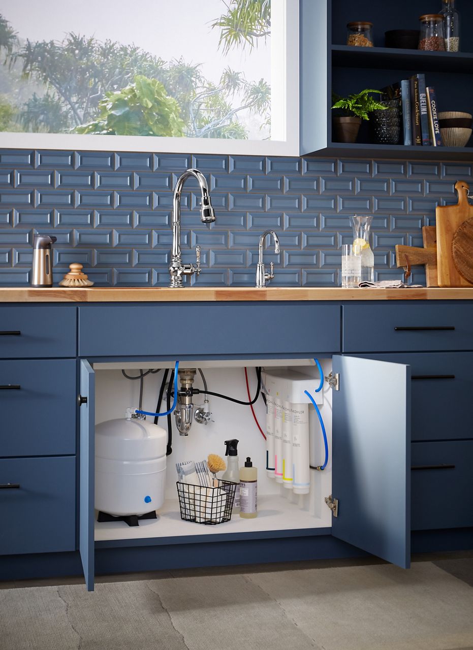 A blue kitchen cabinet with the door below the sink and faucet opened to show a reverse osmosis water filtration system.