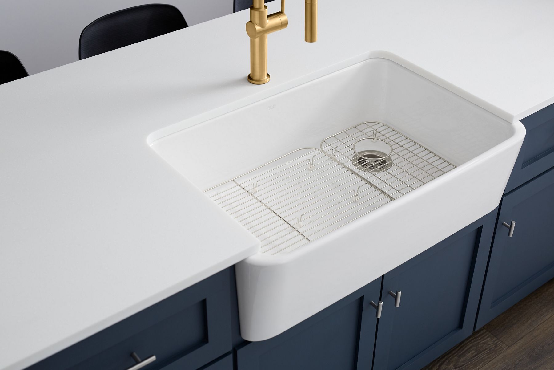 A white farmhouse sink with dark blue cabinets and a brushed moderne brass faucet.