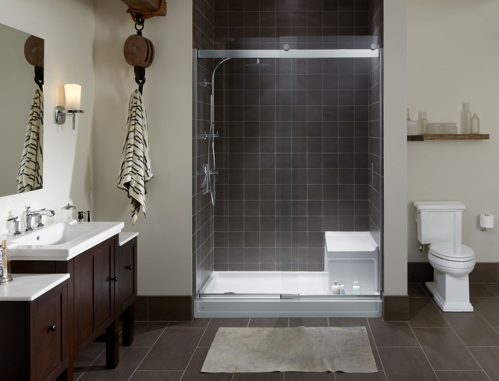 A bathroom with gray square tiled shower with KOHLER Tresham shower base with integral seat, Levity frameless shower doors, shower column, and handshower.