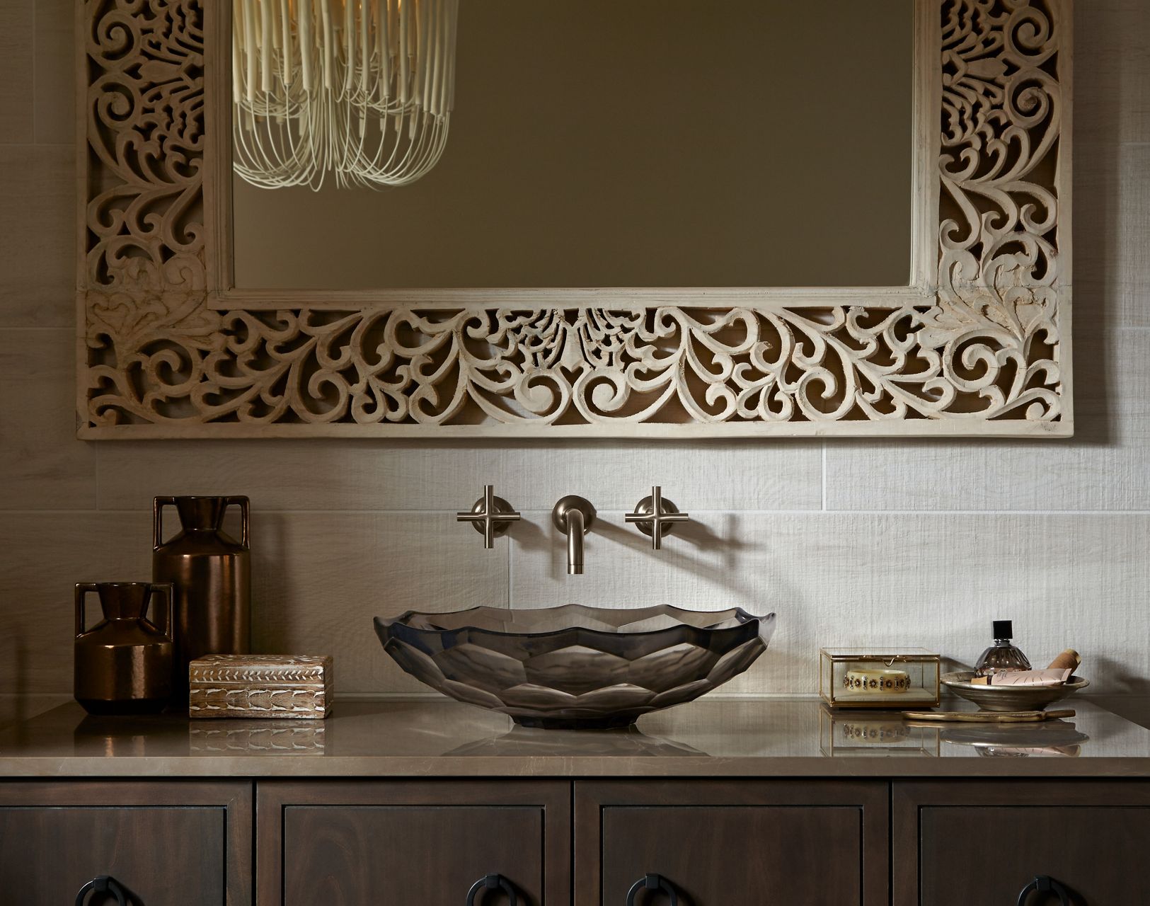 Front view of a dark gray faceted glass Briolette vessel sink on a dark brownish gray vanity with a brushed nickel wall-mount faucet above, and above that an ornamental scroll work piece going horizontally across the wall.