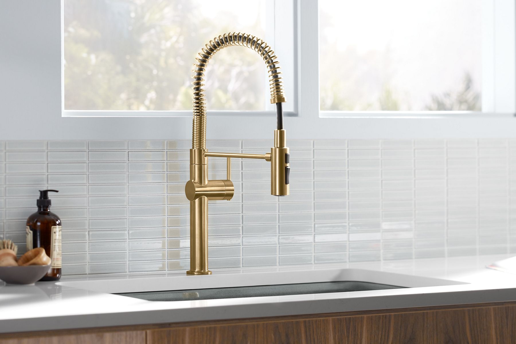 Kohler Crue kitchen sink faucet set in white counter top infront of tiled backsplash with handsoap and supplies to the left.