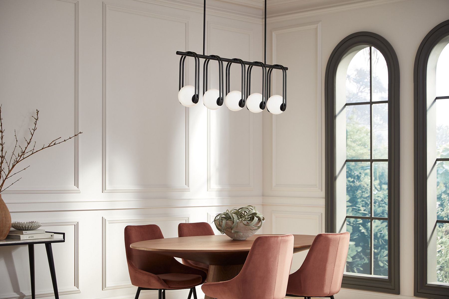 A 5-light KOHLER Kraga chandelier hangs in a white dining room a with large arched window.