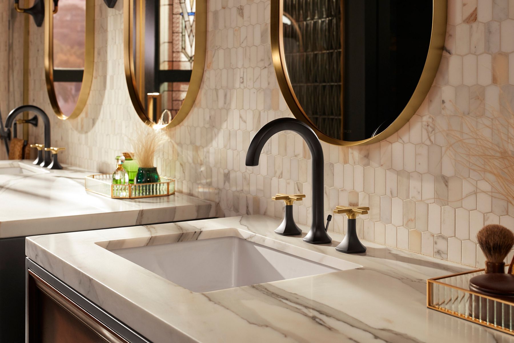 A bathroom vanity space with a stone counter top and tile wall in light cream colors, and a faucet in two tone matte black and brushed brass.
