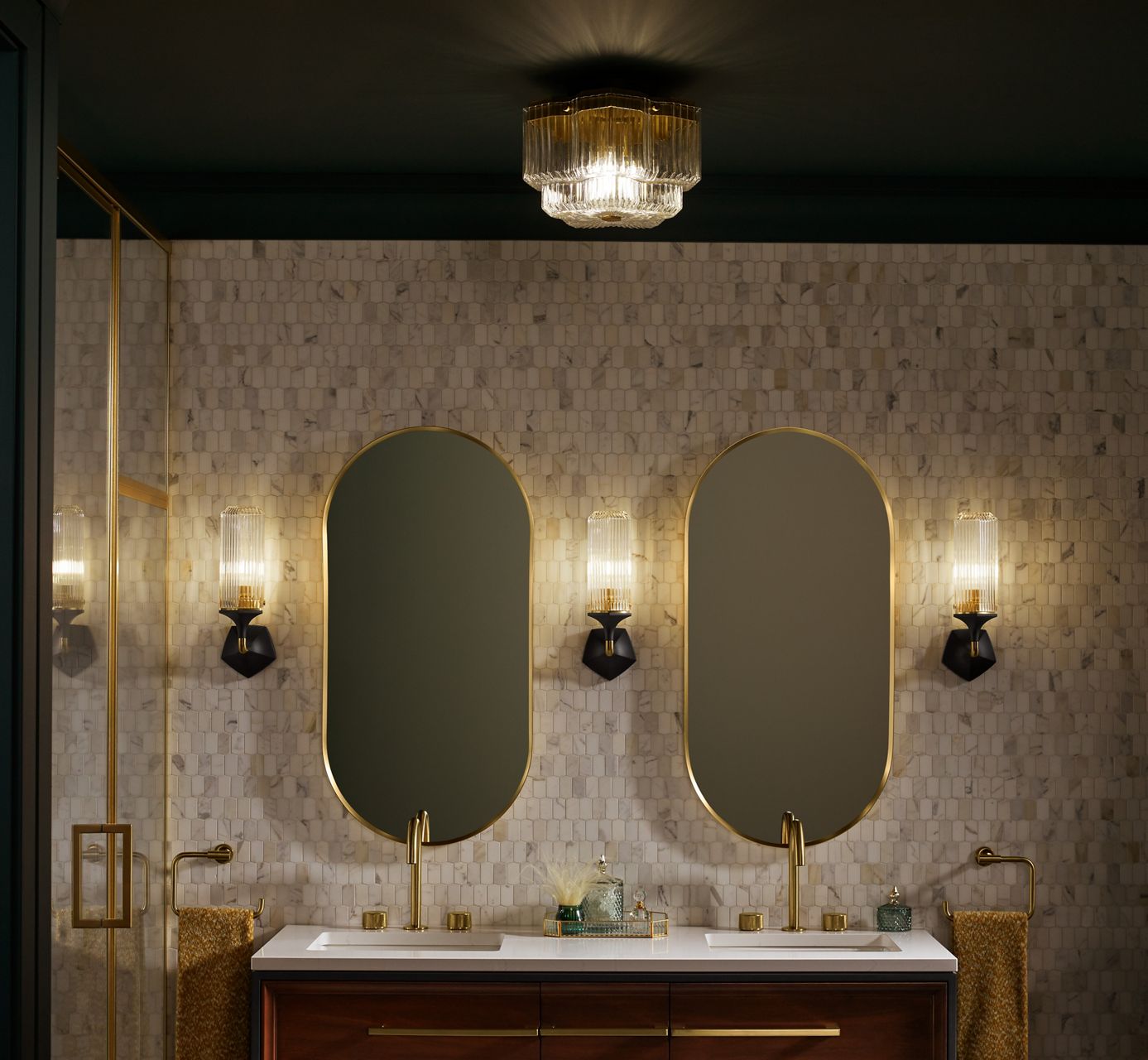 A bathroom with small square stone tiled walls and two capsule mirrors is lit by three KOHLER sconces.