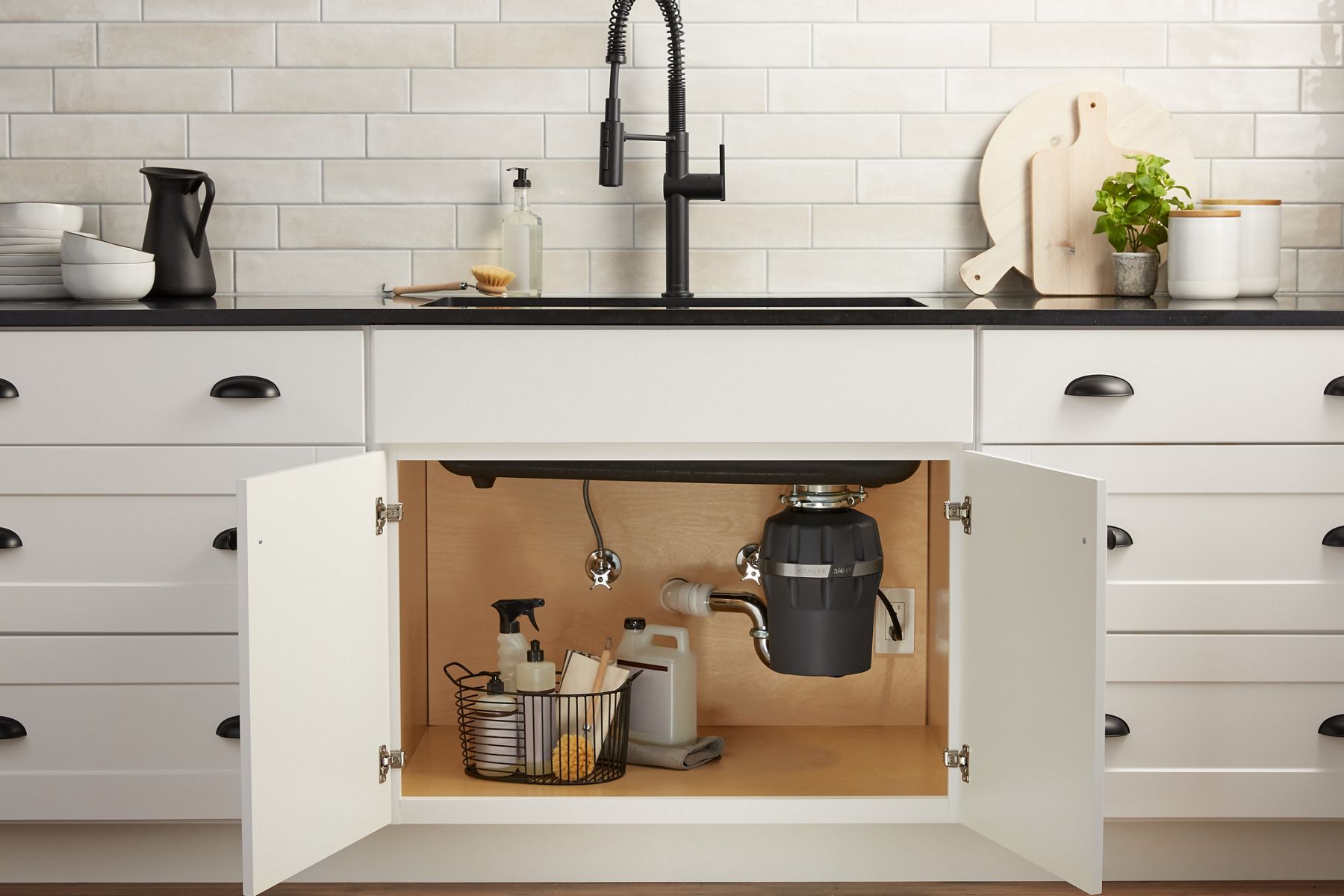 White Shaker style cabinet doors stand open, revealing a KOHLER garbage disposal and a basket of cleaning supplies.
