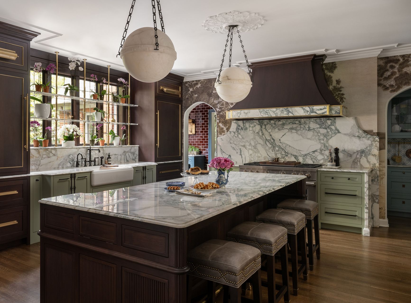 A wide shot lifestyle image of a sunny kitchen elegantly designed with dark wooden cabinets with white and gray marble countertops, gold cabinet hands, bronzed kitchen sink faucets, water tap faucet, and sprayhead, with pale green cabinet accents. There is an island in the center of the image with leather stools. On the island are waffles, pastries, and a vase with pink roses.