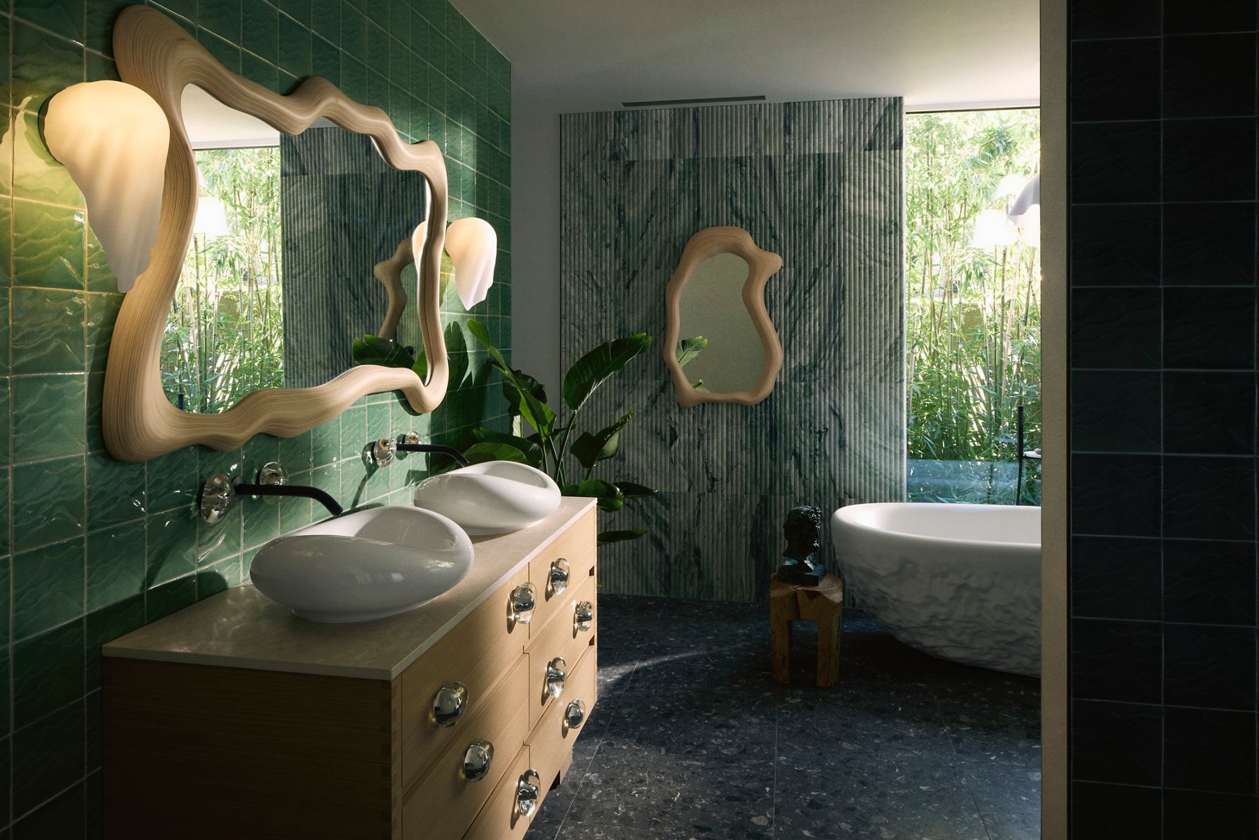A modern bathroom showcasing the Kohler x Arsham Landshapes™ Collection. The image features a white sculptural sink on a wooden vanity, a uniquely shaped wooden mirror, and green tiled walls. In the background, a freestanding white bathtub is partially visible next to large windows that allow natural light to filter in.