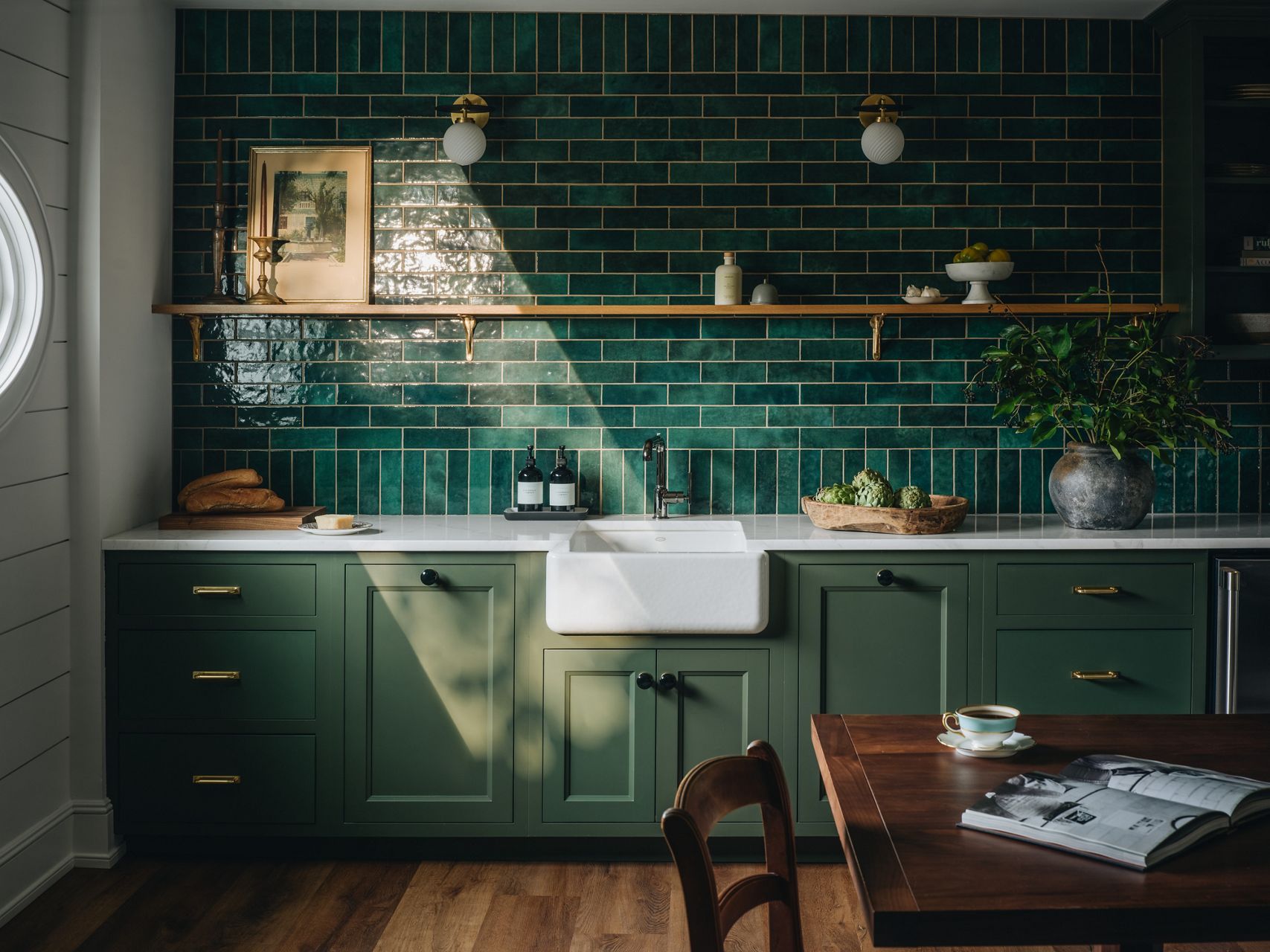 Moody kitchen with deep green cast iron sink, matching green cabinetry, glossy green tile backsplash, open wood shelving, and warm wood flooring.