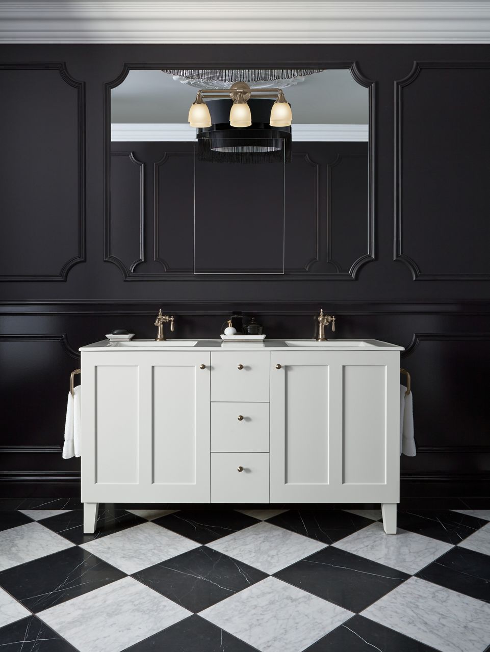 A white KOHLER double vanity with Shaker doors and single handle faucets in a bathroom with black and white checkered floors and dark paneled walls.