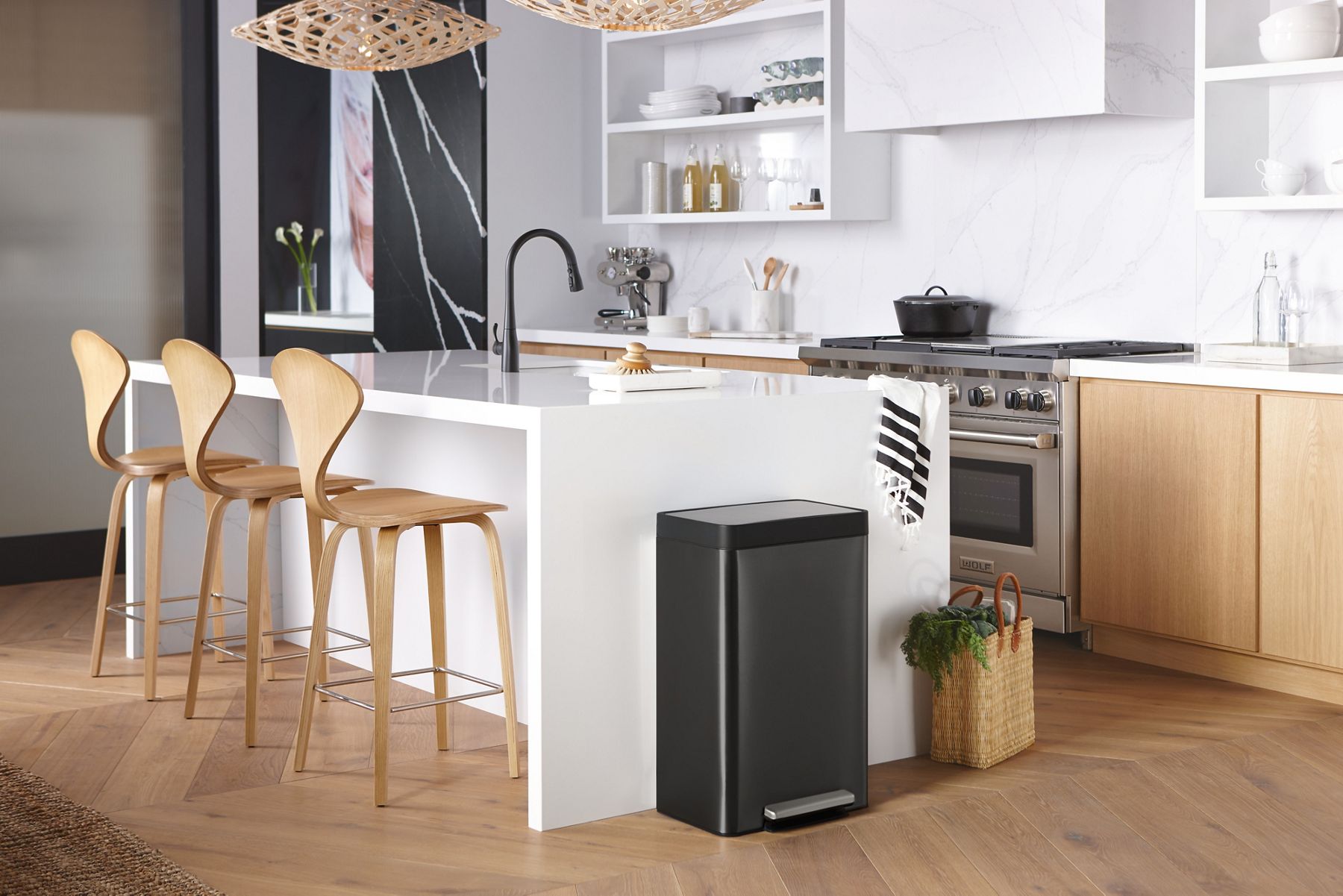 Three wooden stools sit at a white kitchen counter with waterfall edge, and Black Stainless KOHLER Step trashcan at one end.