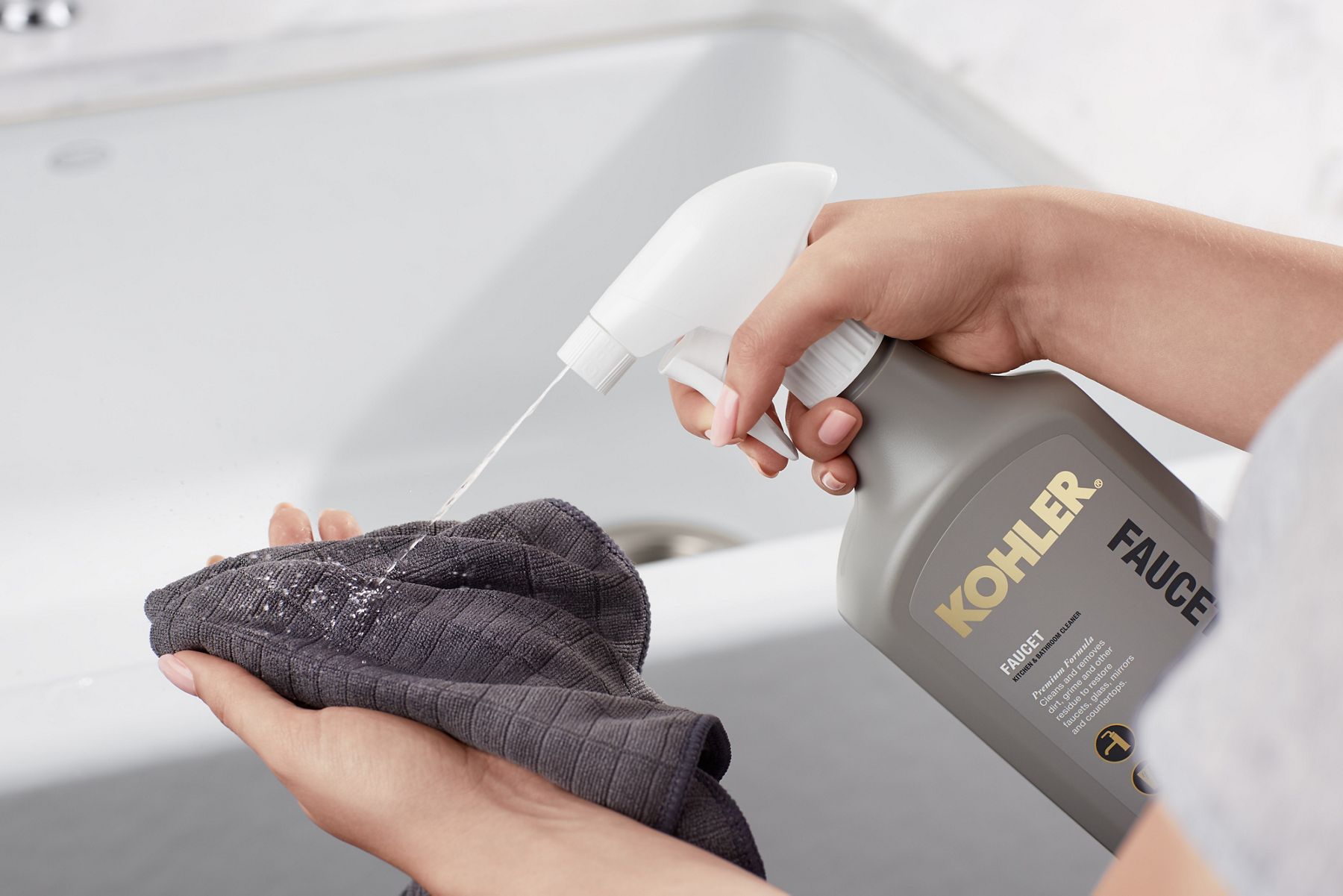 A person sprays KOHLER Faucet cleaner onto a gray cleaning cloth, prior to cleaning their white sink and faucet.