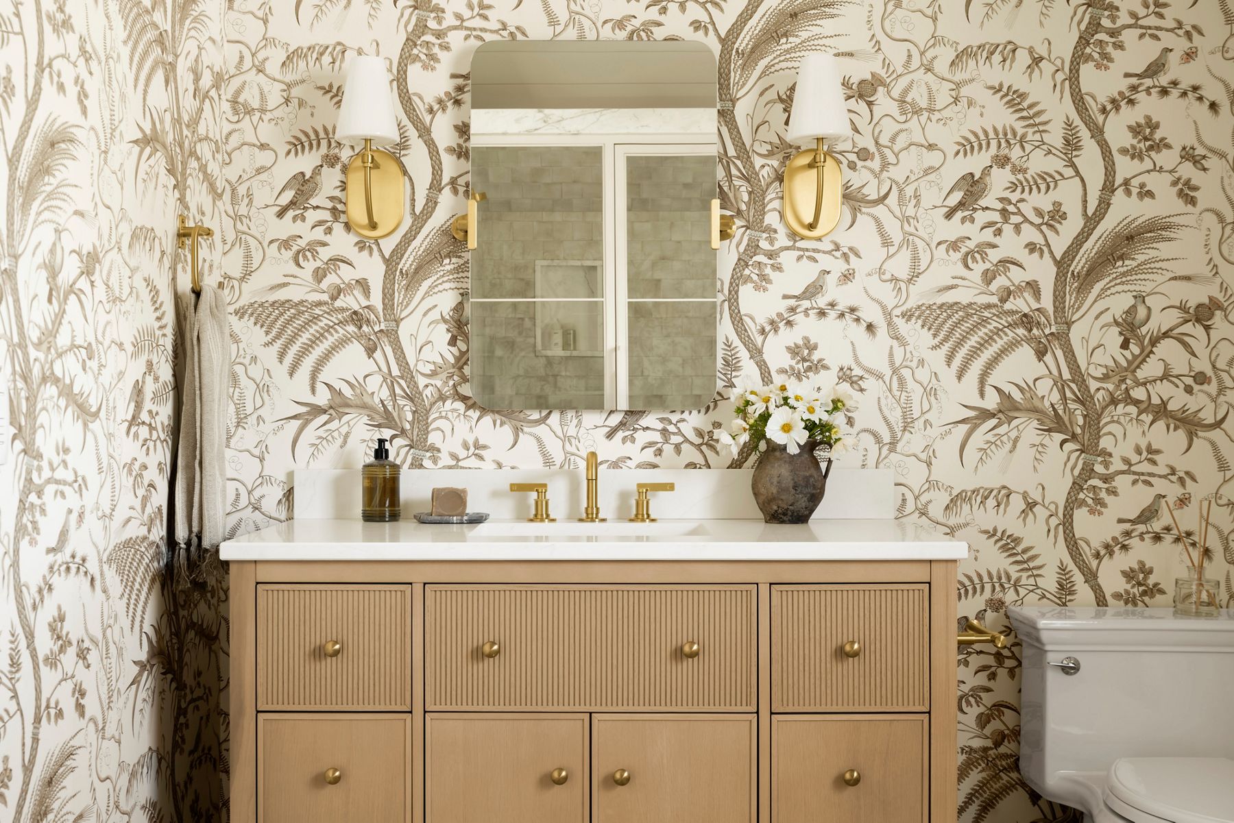 A bathroom vanity in light brown with reeded doors, a white top, and a brushed brass faucet, backed by floral wallpaper in beige and brown tones.