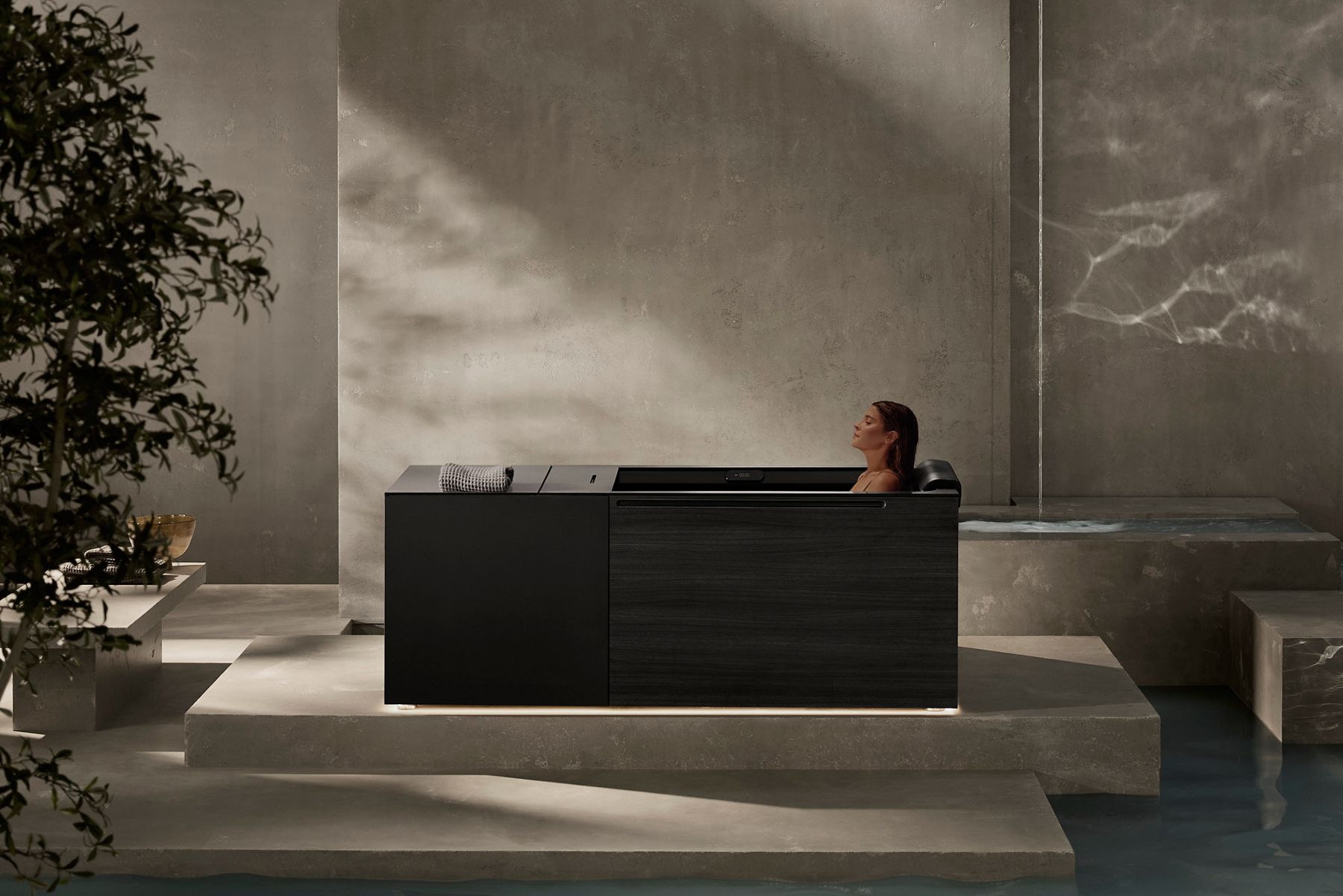 Une salle de bain moderne et minimaliste avec une baignoire à immersion rectangulaire de couleur sombre, la douce lumière du soleil filtrant à travers le mur et une personne qui se détend dans son bain.
