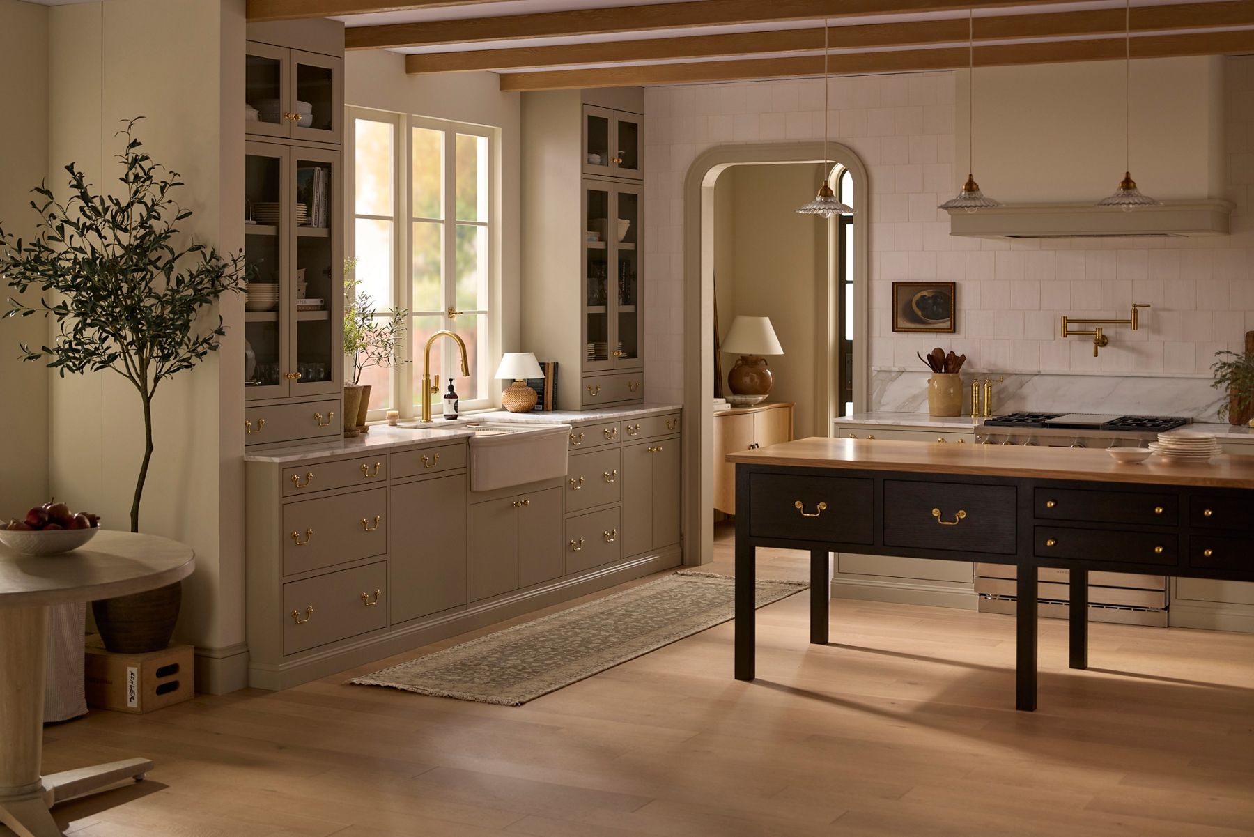 Luxury farmhouse kitchen with green cabinetry, marble countertops, dark wood island, and exposed ceiling beams in natural light