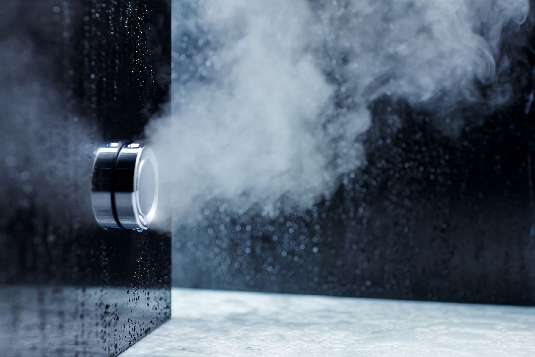 Steam outlet emitting vapor from a chrome steam shower mounted on a wet black glass wall