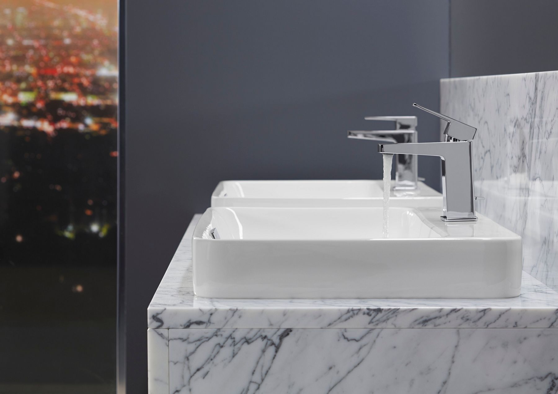 Side view of two white bathroom sinks with chrome faucets in front of a bluish gray wall.