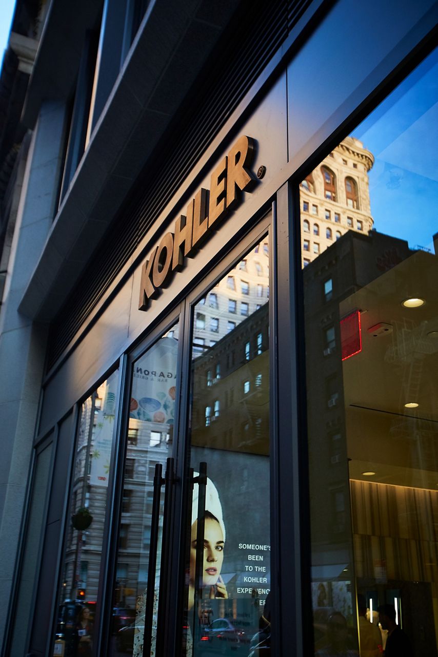 The outside of a KOHLER store, with the name above a doorway that is reflecting the image of nearby buildings.