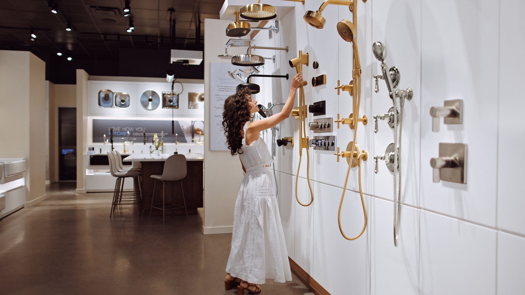 Wide image of a woman standing at the shower display wall in a Kohler Store as she's reaching to pick up a handheld shower from the wall of products. There are more product displays in the background.