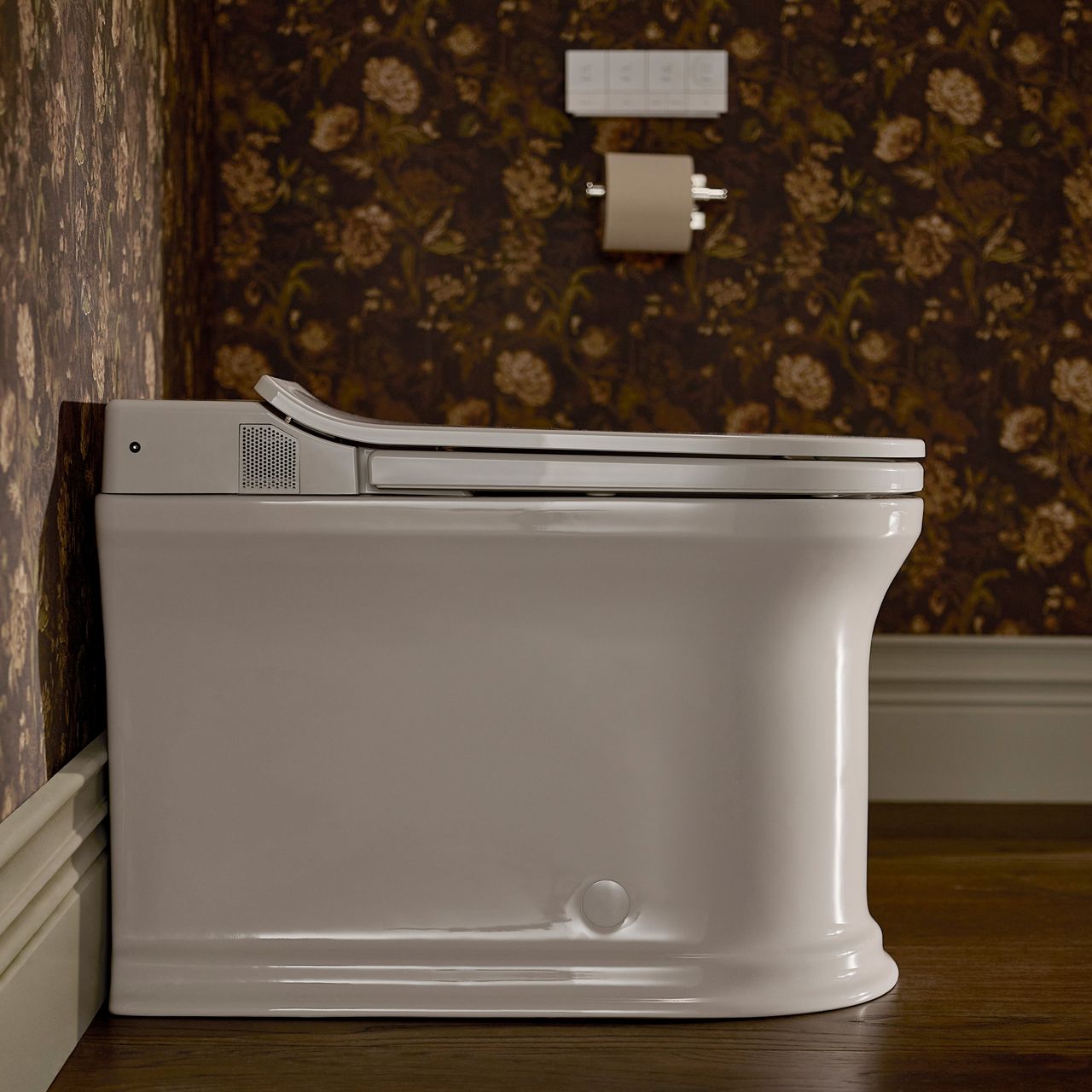 Side view of a smart toilet against a dark floral wallpapered bathroom wall, with a wall-mounted control panel and toilet paper holder above it.
