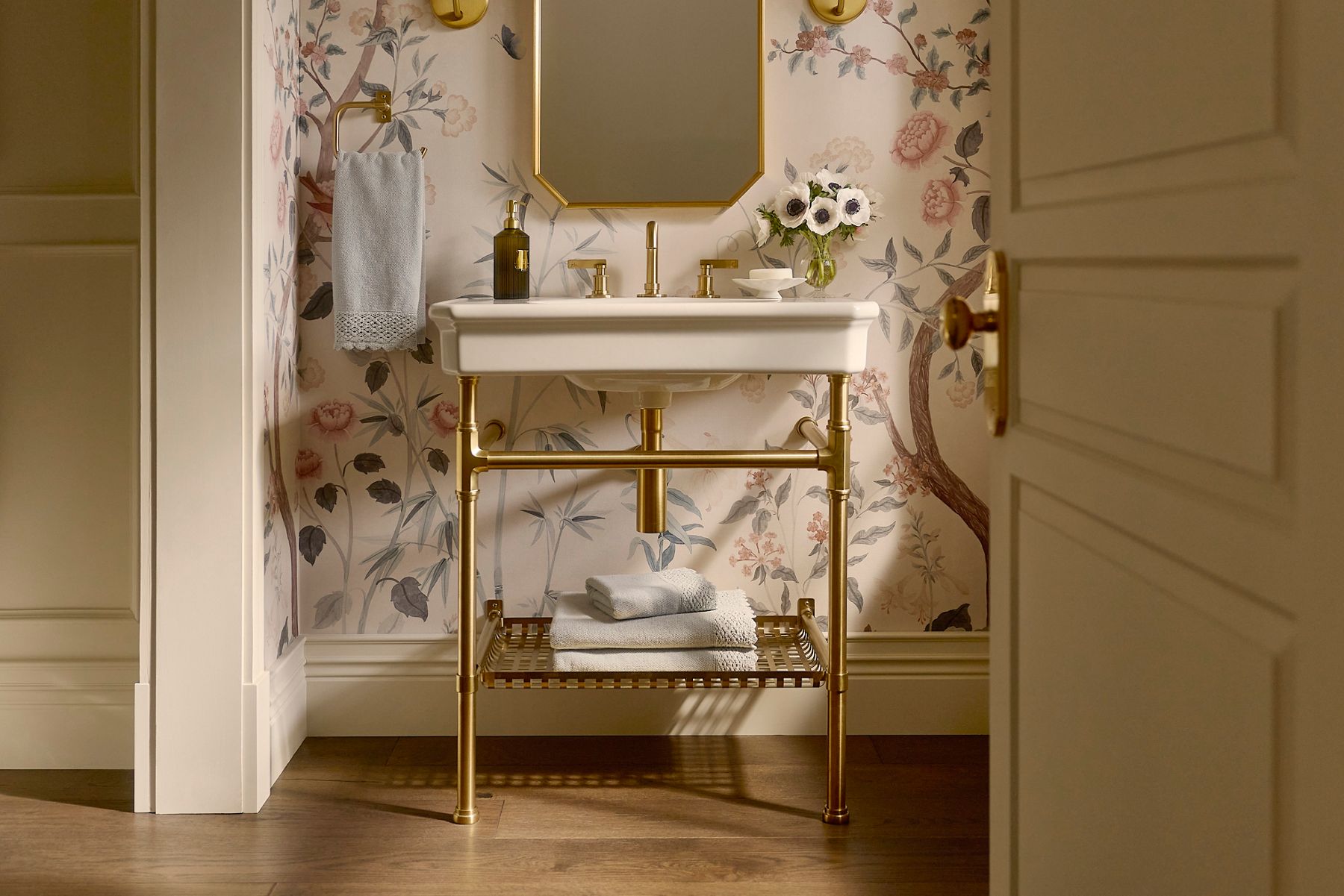 Bathroom console sink with brass legs and fixtures, set against floral wallpaper, featuring a rectangular mirror, a towel hanging to the left, a soap dispenser, and a vase of flowers on the countertop with folded towels on the lower shelf.