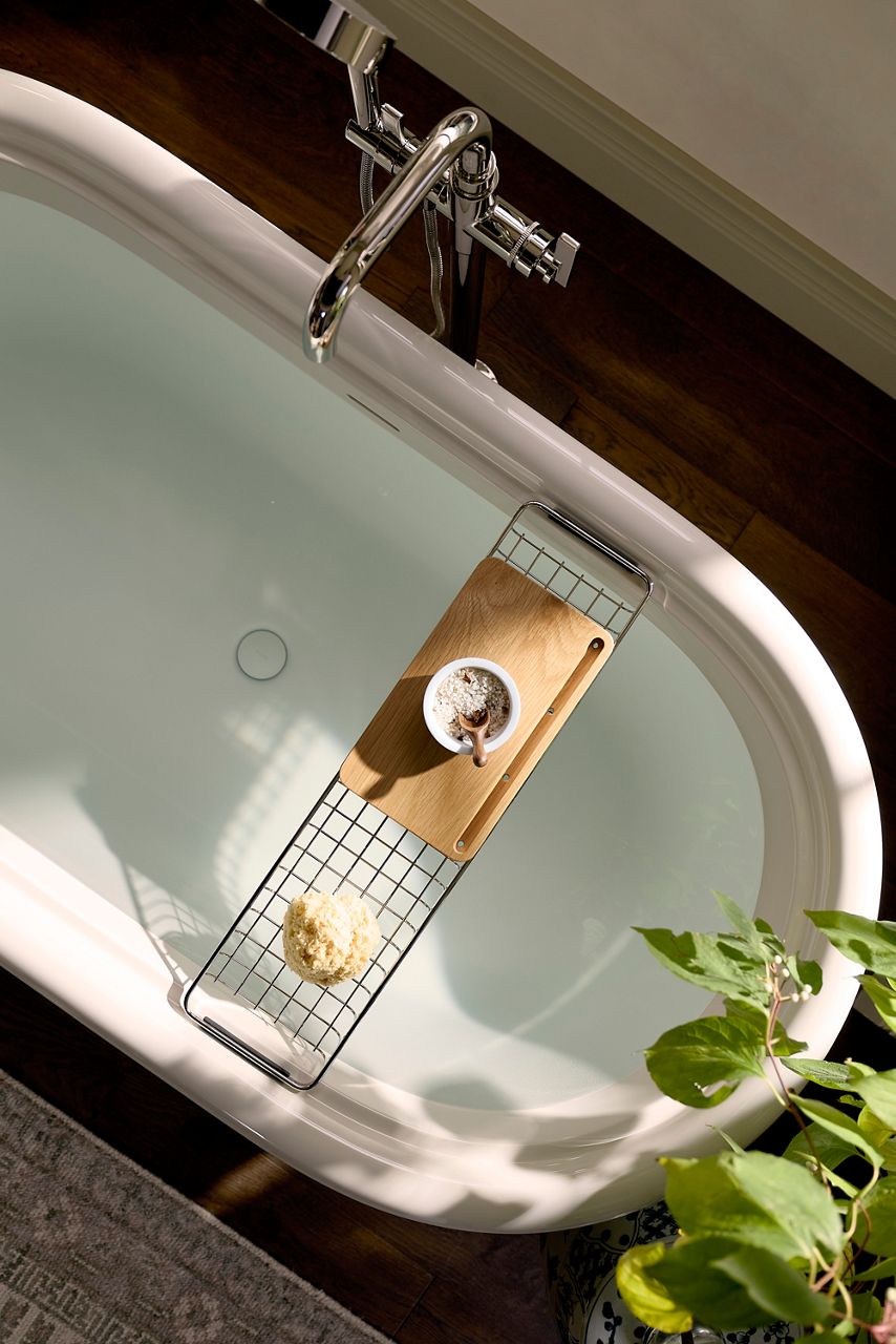 Overhead view of a freestanding bathtub filled with water, featuring a metal and wood bath caddy holding a candle and sponge, with natural light and a green plant beside the tub.
