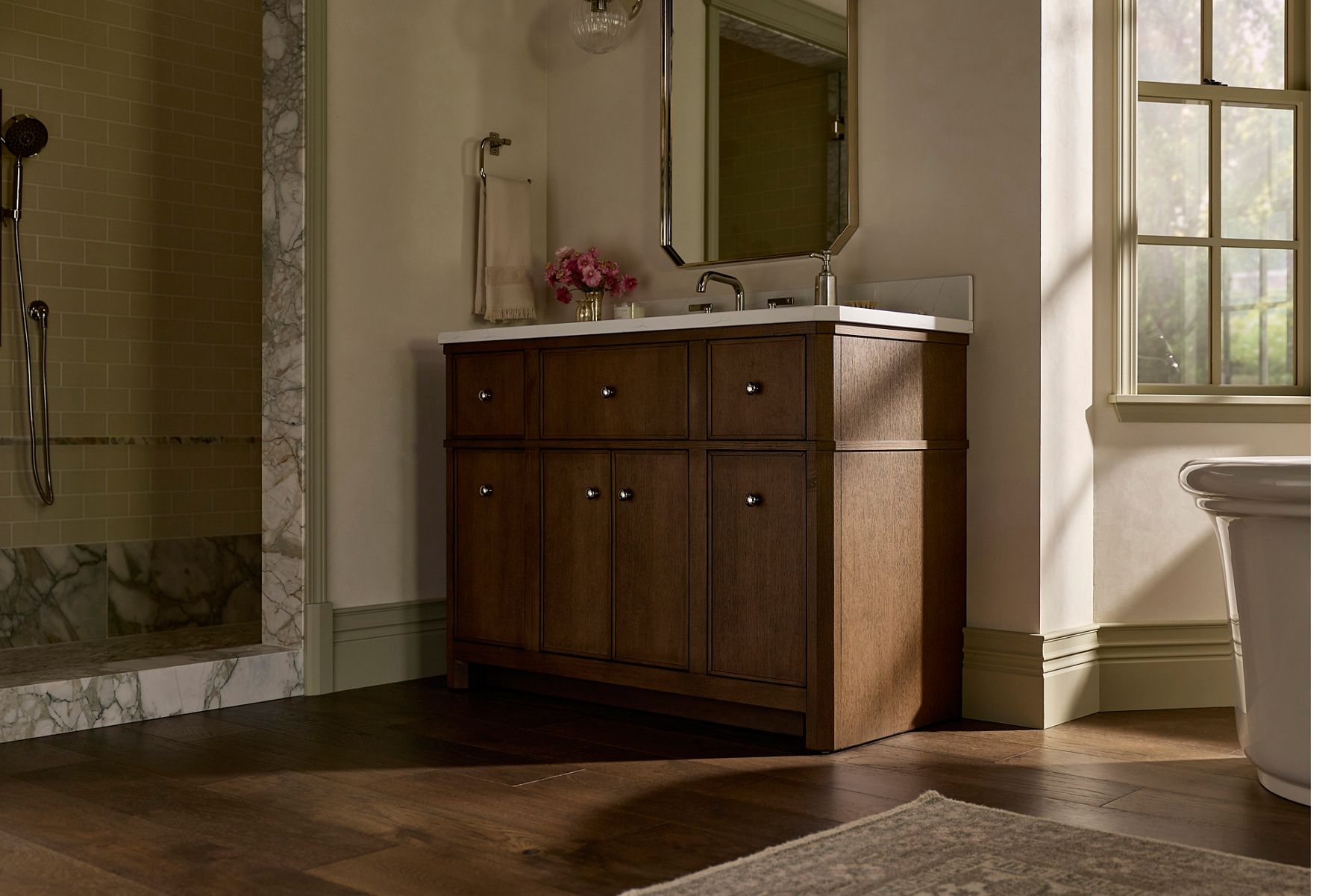 Wood bathroom vanity with multiple drawers and cabinet doors, topped with a white countertop and undermount sink, positioned beneath a framed mirror with a small vase of pink flowers on the counter.