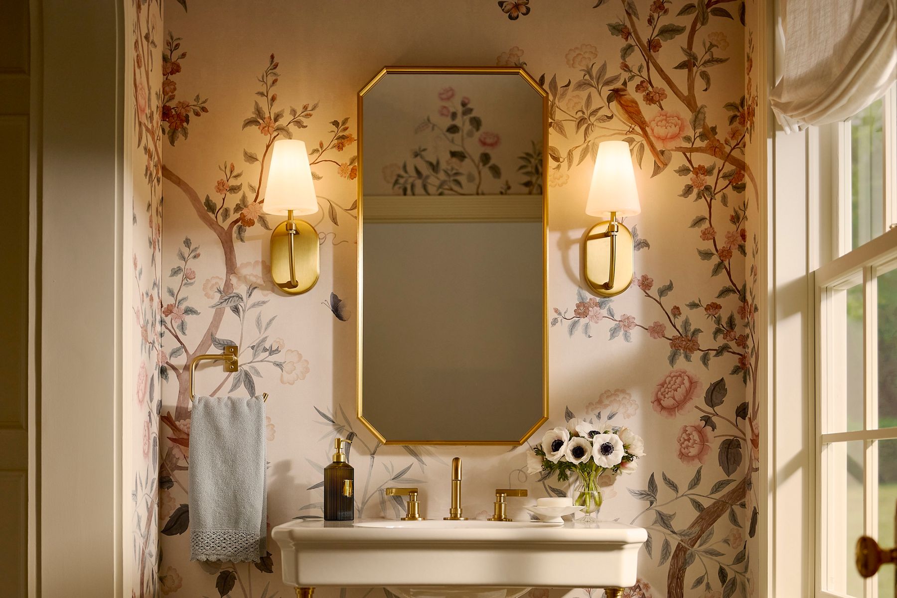 Brass-framed bathroom mirror centered above a sink, flanked by two wall‑mounted sconces with white shades, set against floral wallpaper with a soap dispenser and vase of flowers on the countertop.