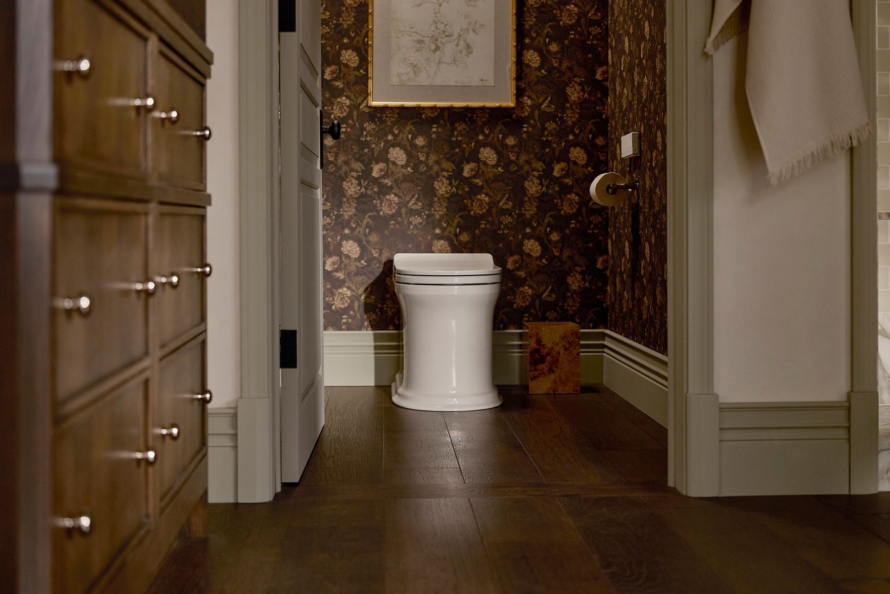 View into the Shea McGee x Kohler Claude primary bathroom featuring a Kohler Claude one-piece elongated smart toilet, dark floral wallpaper, rich wood flooring, and warm wood vanity cabinetry.