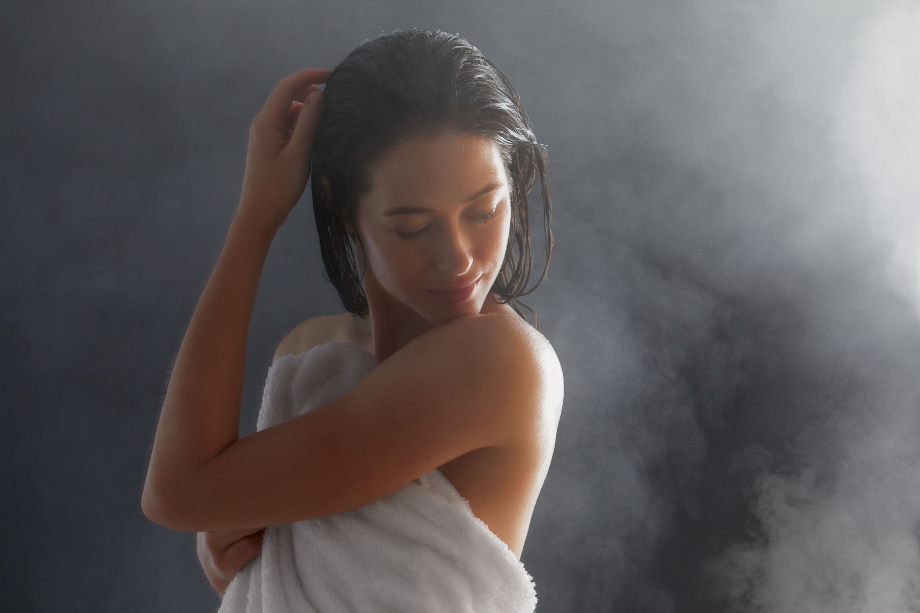 Close-up image of a woman who just exited the shower with wet hair and a towel wrapped around her, as she's looking back at the moisturized skin on her shoulder and upper back.  