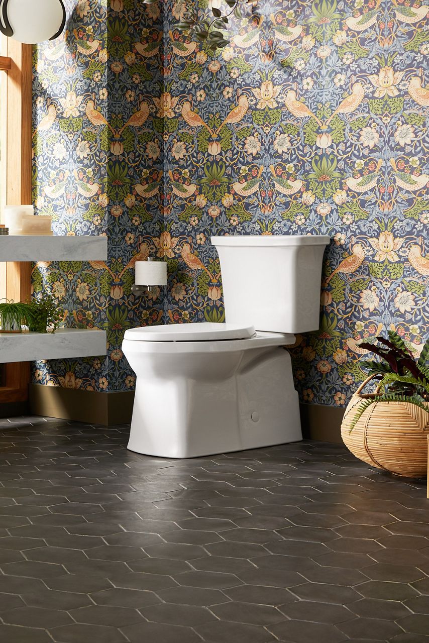 Toilettes modernes blanches dans une salle de bain avec papier peint floral décoratif et carrelage de plancher hexagonal foncé.