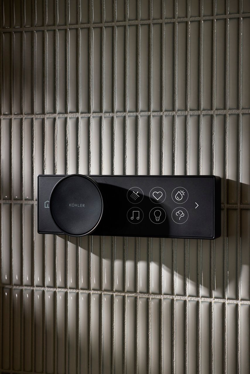 A modern digital control panel mounted on a white tiled wall, featuring a large dial on the left and a series of touch icons on the right, representing various functions such as music, lighting, heart rate, and steam.