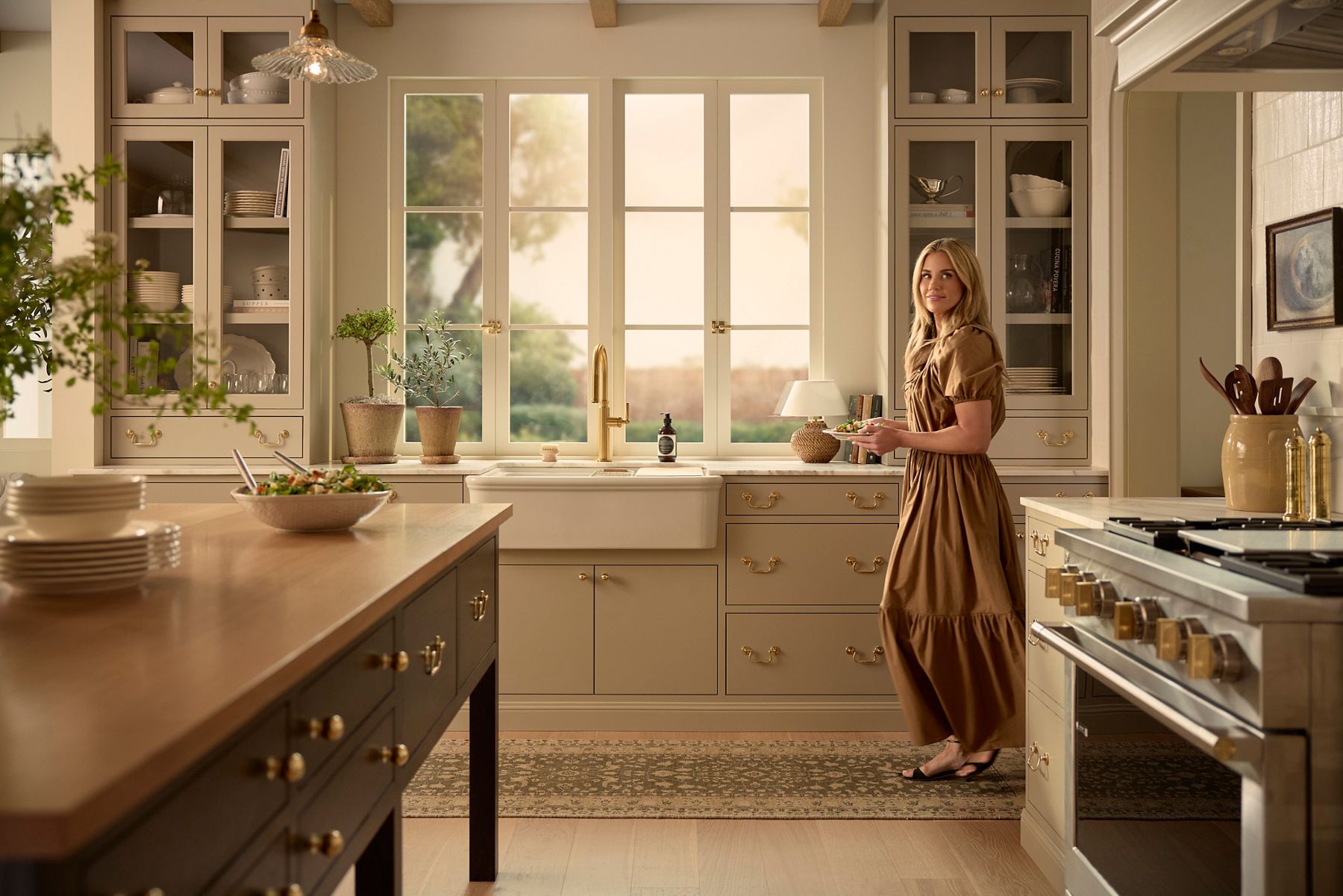 Audrine™ farmhouse sink installed beneath a large window, featuring an apron-front design, tall brass kitchen faucet, and neutral cabinetry with brass hardware.