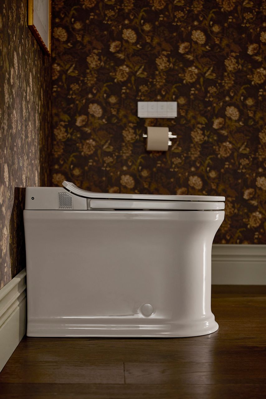 Kohler Claude one-piece elongated smart toilet positioned against dark floral wallpaper in the Shea McGee x Kohler Claude primary bathroom with warm wood flooring and classic trim details.