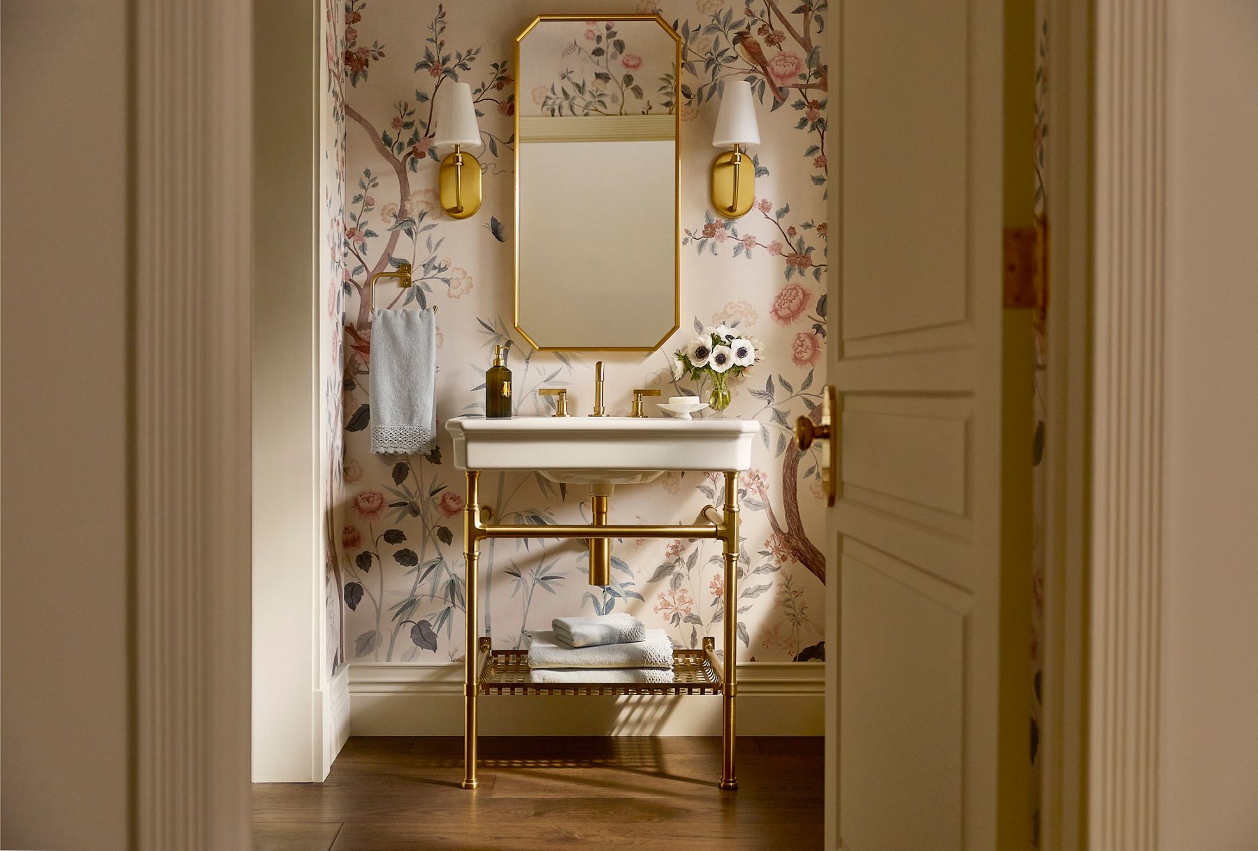 View into the Claude powder room by Shea McGee and Studio McGee featuring floral wallpaper, a white console sink with brass legs and fixtures, an octagonal brass mirror, and coordinating wall sconces.