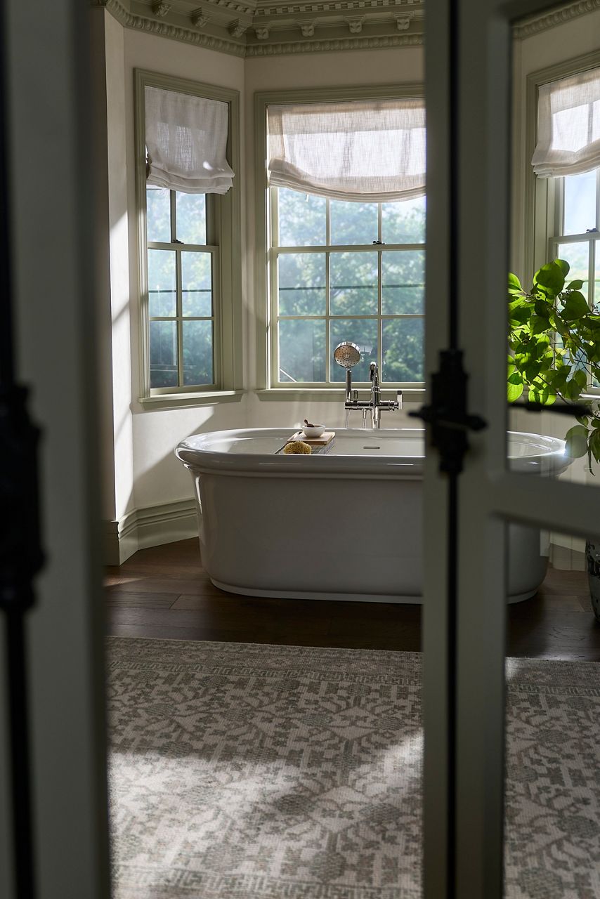 Through partially open doors, a freestanding white bathtub sits in the Shea McGee x Kohler Claude primary bathroom, framed by tall windows, patterned wallpaper, and mosaic tile flooring.