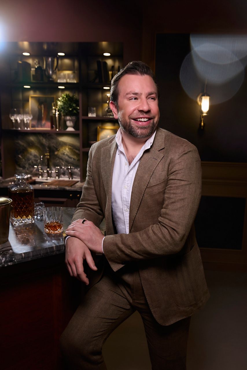 Design expert Javier Burkle leaning against his sophisticated home bar smiling and looking off in the distance.