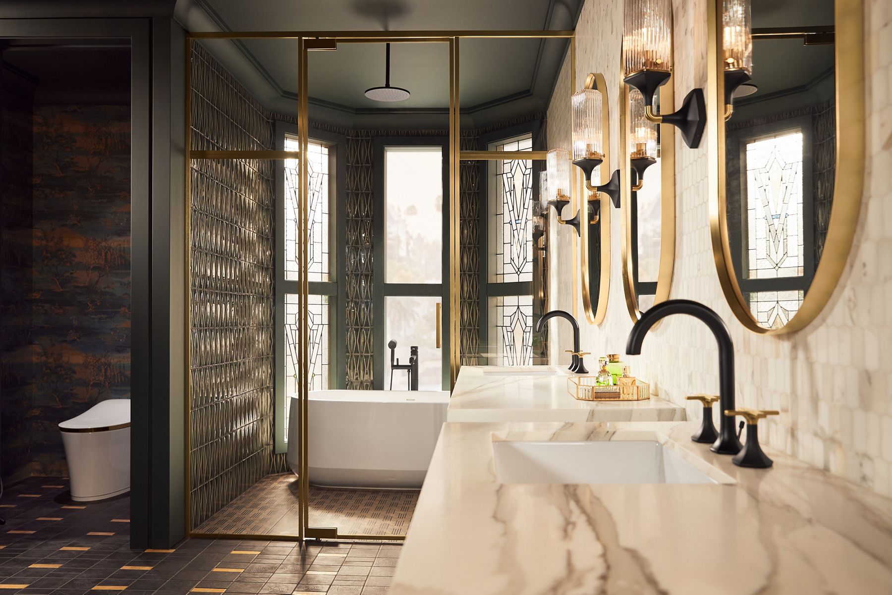 A large, luxurious bathroom looking down the length of two extra-wide single vanities, each of which contains a two-tone faucet, gold-framed mirrors and two-tone sconce lights, beyond which is a brass-framed glassed-in area containing an overhead rainhead, a white freestanding bath and mossy green walls with ornate leaded glass windows.